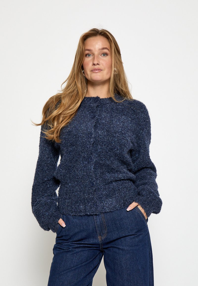 Tumši zila adīta kardigāna ar strukturētu apdari, ar apaļu kakla izgriezumu, pogu aizdari un garām piedurknēm. Valkāts ar tumšiem džinsiem.