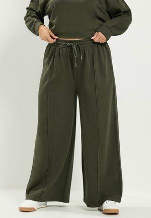 Persona con pantalones anchos de pierna de color verde oscuro y sudadera a juego, ajustándose la cintura, de pie con zapatillas blancas sobre un fondo claro.