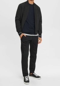 Giacca bomber nera con zip, maglione a girocollo blu navy, pantaloni neri e sneakers nere con lacci bianchi. Design casual e aderente.