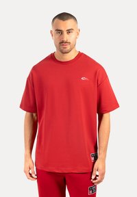 Camiseta de algodón roja con mangas cortas, escote redondo y un logo blanco en el pecho. Presenta una etiqueta en el dobladillo.