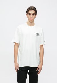 Λευκό βαμβακερό t-shirt με κοντά μανίκια, στρογγυλή λαιμόκοψη, που διαθέτει μαύρο τυπωμένο λογότυπο στο αριστερό στήθος και μαύρη λεπτομέρεια στο αριστερό μανίκι.