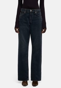 Donkerblauwe, high-waist jeans met wijde pijpen, voorzien van het klassieke ontwerp met vijf zakken, metalen knoopsluiting en subtiele vervaging, gedragen door een model.