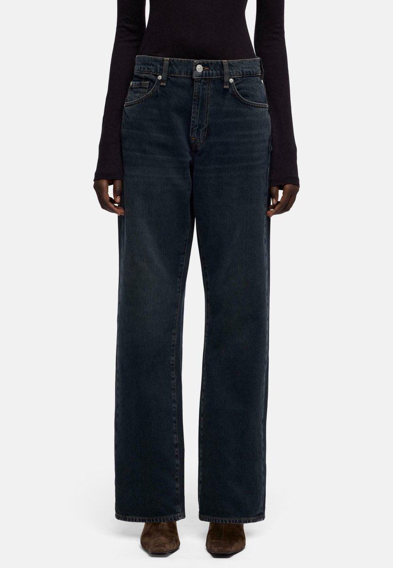 Donkerblauwe, high-waist jeans met wijde pijpen, voorzien van het klassieke ontwerp met vijf zakken, metalen knoopsluiting en subtiele vervaging, gedragen door een model.