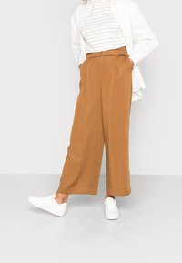 Henkilö, jolla on päällään korkeavyötäröiset laajat ruskeat housut, valkoiset lenkkarit, raidallinen paita ja auki oleva valkoinen nappipaita, seisoo valkoisella lattialla.