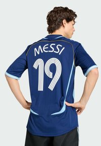 Persona che indossa una maglia da calcio blu con "MESSI" e il numero "19" stampati sul retro, in piedi con le mani sui fianchi.