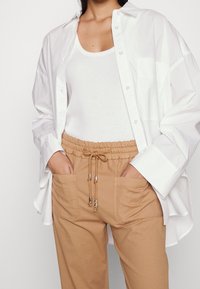 White button-up shirt over a white tank top, paired with beige drawstring pants featuring side pockets and silver hardware accents.