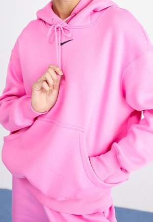 Personne portant un sweat à capuche rose vif avec un logo Nike noir, tirant sur un cordon de serrage et l'autre main dans la poche avant.