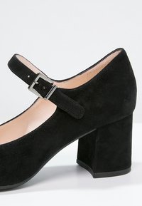 Chaussure Mary Jane en suede noir avec un bout arrondi, un talon bloc haut et une sangle ajustable ornée d'une boucle en argent. Doublure intérieure lisse.