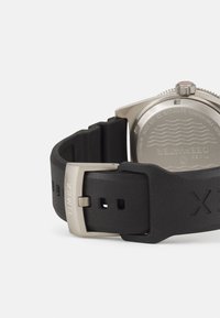 Bracelet de montre en caoutchouc noir avec une boucle en métal argenté. Le fond du boîtier en acier inoxydable présente un motif de vagues en relief et une gravure "Timex".