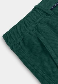 Donkergroen textiel met textuur, elastische tailleband en zichtbare zijnnaad, voorzien van een klein zwart label met witte tekst.