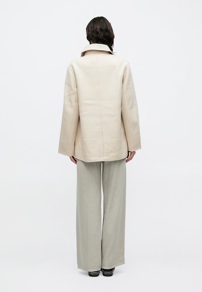 Bézs színű, túlméretezett zakó strukturált gallérral, egyenes szabással és texturált anyagból, világosszürke, széles szárú nadrággal párosítva.