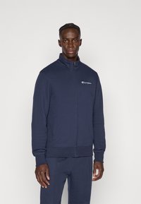 Φούτερ σε ναυτικό μπλε χρώμα με φερμουάρ, κατασκευασμένο από μαλακό ύφασμα και με ψηλό γιακά, διαθέτει μικρό λευκό λογότυπο στο αριστερό στήθος και πλαϊνές τσέπες.