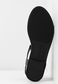 Semelle de sandale plate noire avec une bande de roulement texturée et le logo "Tommy Hilfiger", appuyée contre un coin blanc.