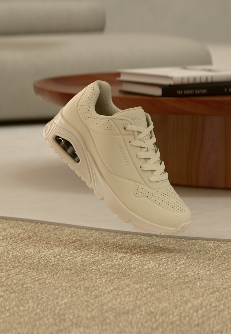 Μπεζ αθλητικό παπούτσι με πλέγμα στην επάνω όψη, κλείσιμο με κορδόνια και διάτρητα διακοσμητικά. Δια θέτει ορατή μονάδα αερόσακου στην σόλα.