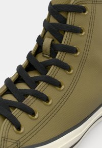 Sneaker înalt de pânză culoare verde măslin cu șireturi și ochiuri negre. Suprafață texturată, talpă din cauciuc și detalii de cusătură întărită.