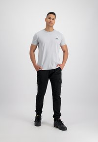 Svetlo siva majica s kratkimi rokavi in okroglim izrezom z majhnim logotipom, v kombinaciji s črnimi cargo hlačami, ki imajo stranske žepe, in črnimi supergami. Čist, sproščen videz.