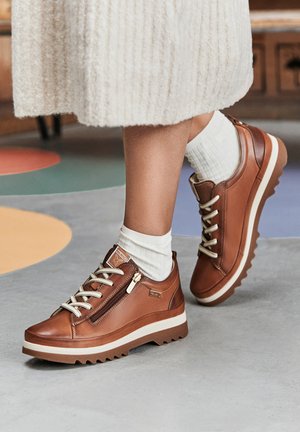 Brown leather sneakers feature a textured design, contrasting cream sole, lace and zipper closures, and a chunky, ridged outsole.