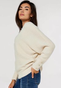 Sweter w kolorze kremowym, prążkowany, o luźnym kroju, z szerokim dekoltem i guzikami na rękawach. W połączeniu z dżinsami.