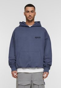 Blue oversized hoodie made from soft fabric, featuring a front pocket and a black printed logo on the chest. Underlayer is a white shirt.