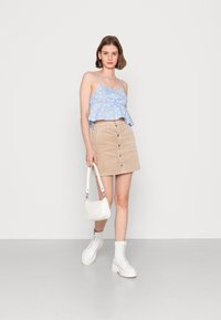 Top curto floral azul com alças ajustáveis, combinado com uma saia de veludo corduroy bege com botões à frente. A modelo segura uma pequena handbag branca e usa botas brancas.