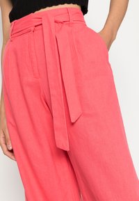Calças cor-de-rosa coral com um cinto de laço, feitas de um tecido leve. Apresentam bolsos laterais e um corte relaxado com detalhes de costura visíveis.