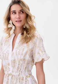 Witte jurk met gouden bloemenborduursel, korte mouwen met een gekartelde rand, V-halslijn en een elastische tailleband. Lang, golvend haar.