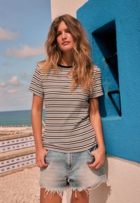 Camiseta de rayas en material suave con mangas cortas y ribete negro en el cuello; combinada con shorts de mezclilla claros con bordes deshilachados, sobre un fondo de pared azul.