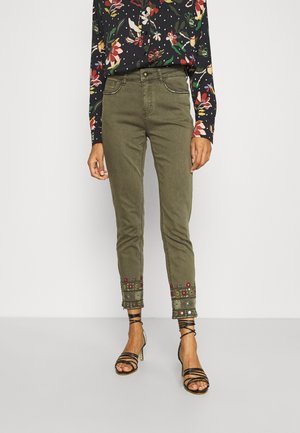 Vrouw draagt olijfgroene korte broek met decoratieve boorden, bloemenprint blouse met lange mouwen, en zwarte sandalen met hoge hak en bandjes.
