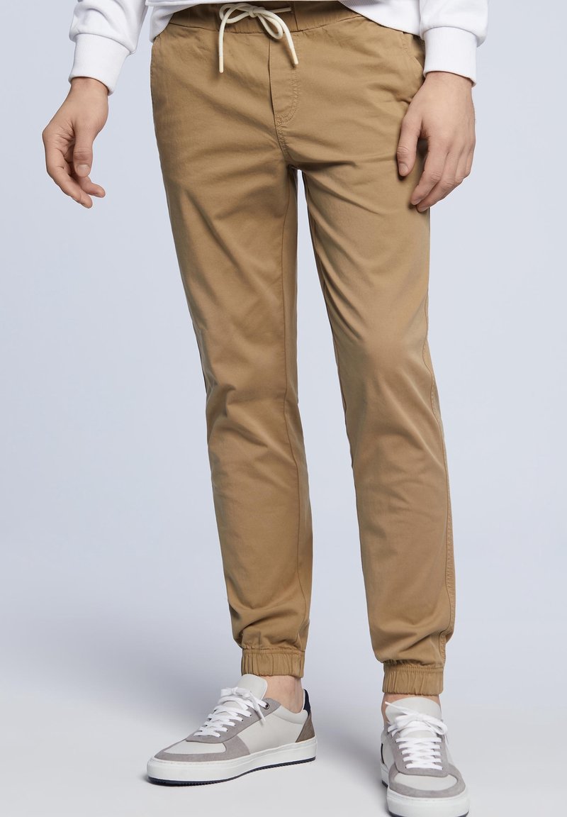 Homme portant un pantalon de jogging à cordon de serrage marron clair et des baskets blanches, debout devant un fond clair uni.