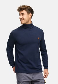 Pull col roulé bleu marine avec texture côtelée, manches longues et un petit patch logo marron sur la poitrine, fabriqué à partir d'un matériau doux.
