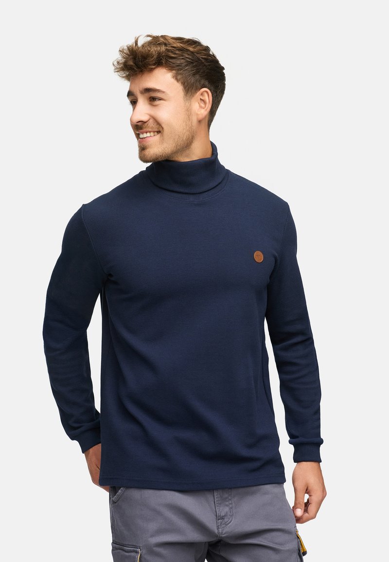 Pull col roulé bleu marine avec texture côtelée, manches longues et un petit patch logo marron sur la poitrine, fabriqué à partir d'un matériau doux.