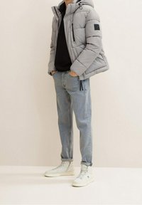 Szara puchowa kurtka z kapturem, kieszeniami na zamek, czarnymi akcentami, w zestawie z jasnoniebieskimi dżinsami i białymi wysokimi trampkami. Gładka faktura.