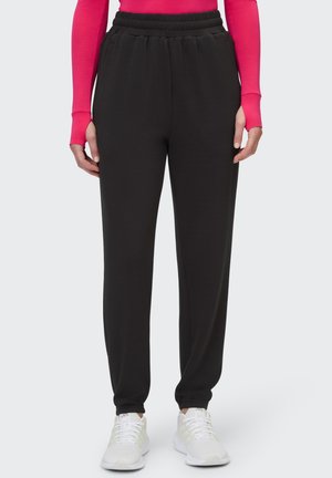 Person trägt schwarze Jogginghosen mit elastischem Bund, weiße Sneakers und ein langärmeliges rosa Oberteil mit Daumenlöchern vor hellem Hintergrund.