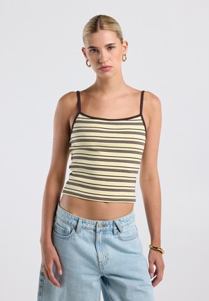 Jonge vrouw met blond haar draagt een beige en bruin gestreepte tanktop, lichtblauwe high waist jeans, gouden creolen en een gouden armband.