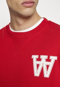 Červená mikina s bílým kulatým výstřihem, s velkým bílým vyšitým logem písmene "W" na hrudi. Hladká textura, volnočasový design.