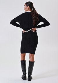 Vrouw met lang donker haar in een lage paardenstaart, gekleed in een zwarte kabelgebreide trui en rok, zwarte kniehoge laarzen, staand met handen op de heupen.