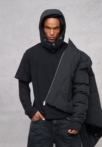 Giubbotto imbottito nero con cappuccio e bottoni a scatto, indossato sopra una maglietta nera. Presenta una chiusura con zip e polsini a costine. Texture morbida e opaca.
