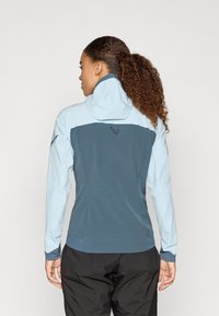 Mėlyna ir žalia striukė, su prigludusia forma, tekstūruota medžiaga ir gobtuvu. Galinėje pusėje yra logotipas, o rankovės kontrastuoja. Juodos kelnės.