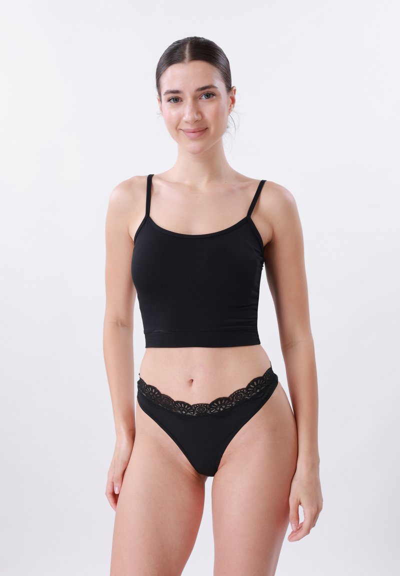 Jeune femme souriante portant un débardeur court noir et une lingerie noire assortie bordée de dentelle sur un fond uni.