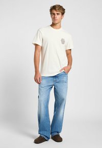 Lichtgekleurde katoenen T-shirt met korte mouwen en een borstzak met een bedrukt logo. Gecombineerd met lichtblauwe wijde jeans en bruine schoenen.