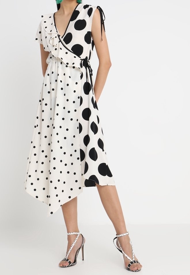 Femme portant une robe blanche asymétrique avec des pois noirs mélangés, grands et petits, associée à des sandales à talons assorties.