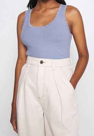 Frau trägt ein geripptes, hellviolettes Tanktop mit Spitzenbesatz und hoch taillierte, lockere, cremefarbene Hose mit einer Hand in der Tasche.