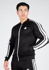 Veste zippée de sport noire avec des bandes blanches sur les côtés, manches longues et un logo sur la poitrine. Présente un design ajusté et des poches avant.