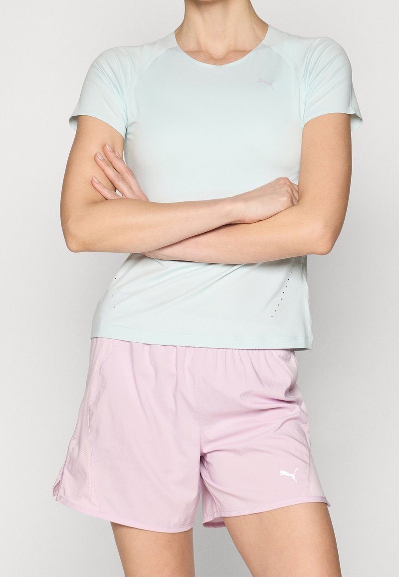 Henkilö, joka on pukeutunut vaaleansinisessä lyhythihaisessa urheilupaidassa ja vaaleanpunaisissa Puma-shorteissa, seisoo käsivarret ristissä tasaisella taustalla.