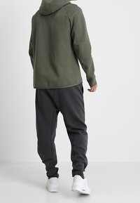 Mörkgrön huvtröja med dragsko och raglanärmar, kombinerad med mörkgrå avsmalnande joggers och vita sneakers. Slät tygstruktur.