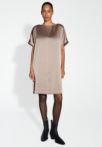 Tailliertes satin taupe Kleid mit einem Boot-Ausschnitt, kurzen Ärmeln und schlichter Silhouette. Kombiniert mit schwarzen Knöchel-Stiefeln und transparenten schwarzen Strumpfhosen.