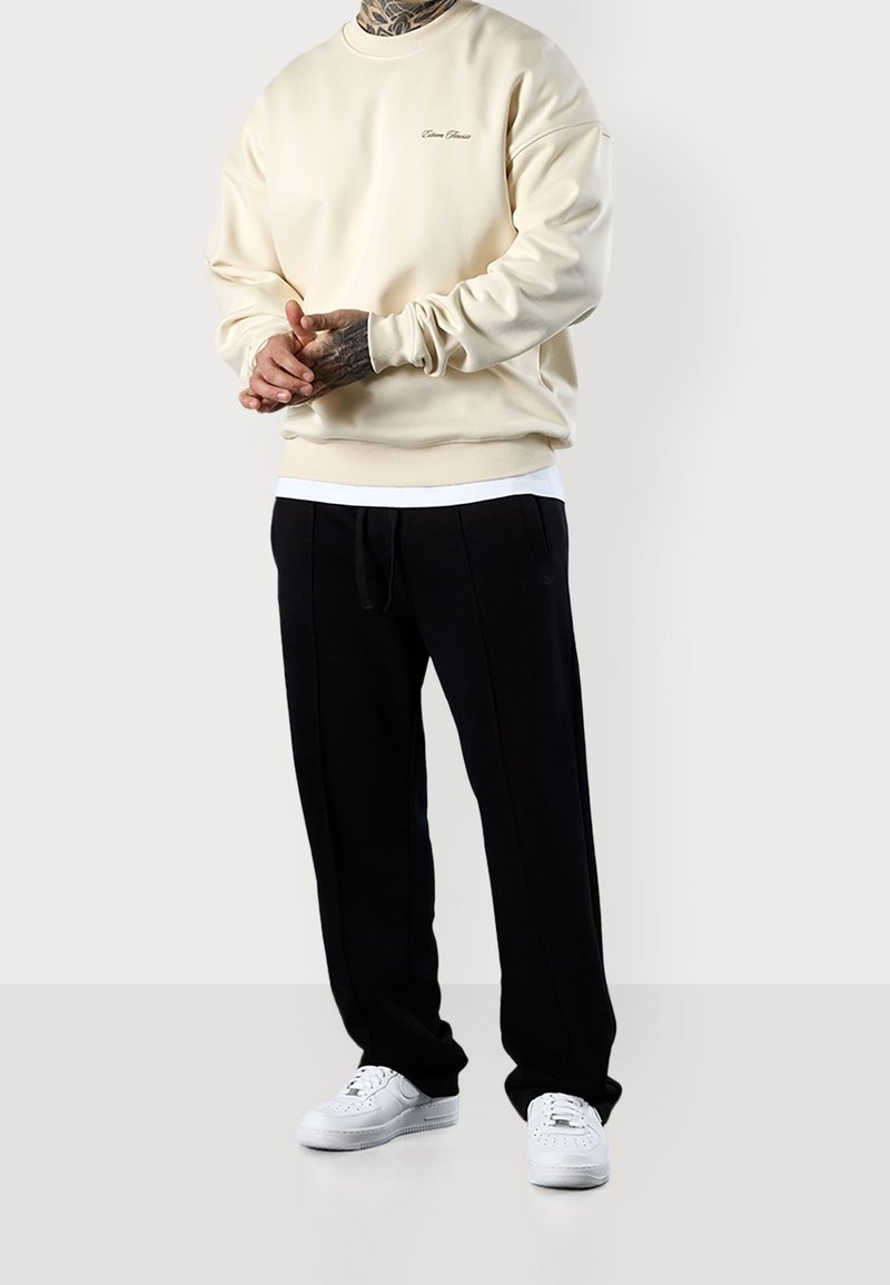 Felpa beige con colletto a giro e logo ricamato, abbinata a pantaloni jogger neri con vita regolabile e tasche laterali, indossata con sneakers bianche.