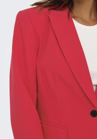 Blazer rouge ajusté avec revers standard, fermeture à un bouton, et poches latérales visibles. Texture de tissu lisse et design cintré.