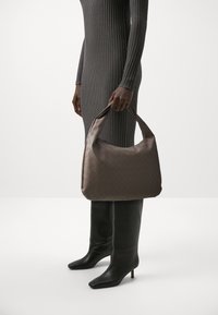 Brązowa, pleciona torebka ze skóry z wyraźnie zarysowanym kształtem, z okrągłym uchwytem. Modelka nosi szarą sukienkę o prążkowanej fakturze oraz czarne kozaki do kolan.