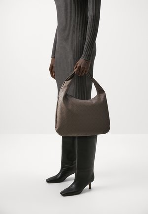 Brązowa, pleciona torebka ze skóry z wyraźnie zarysowanym kształtem, z okrągłym uchwytem. Modelka nosi szarą sukienkę o prążkowanej fakturze oraz czarne kozaki do kolan.